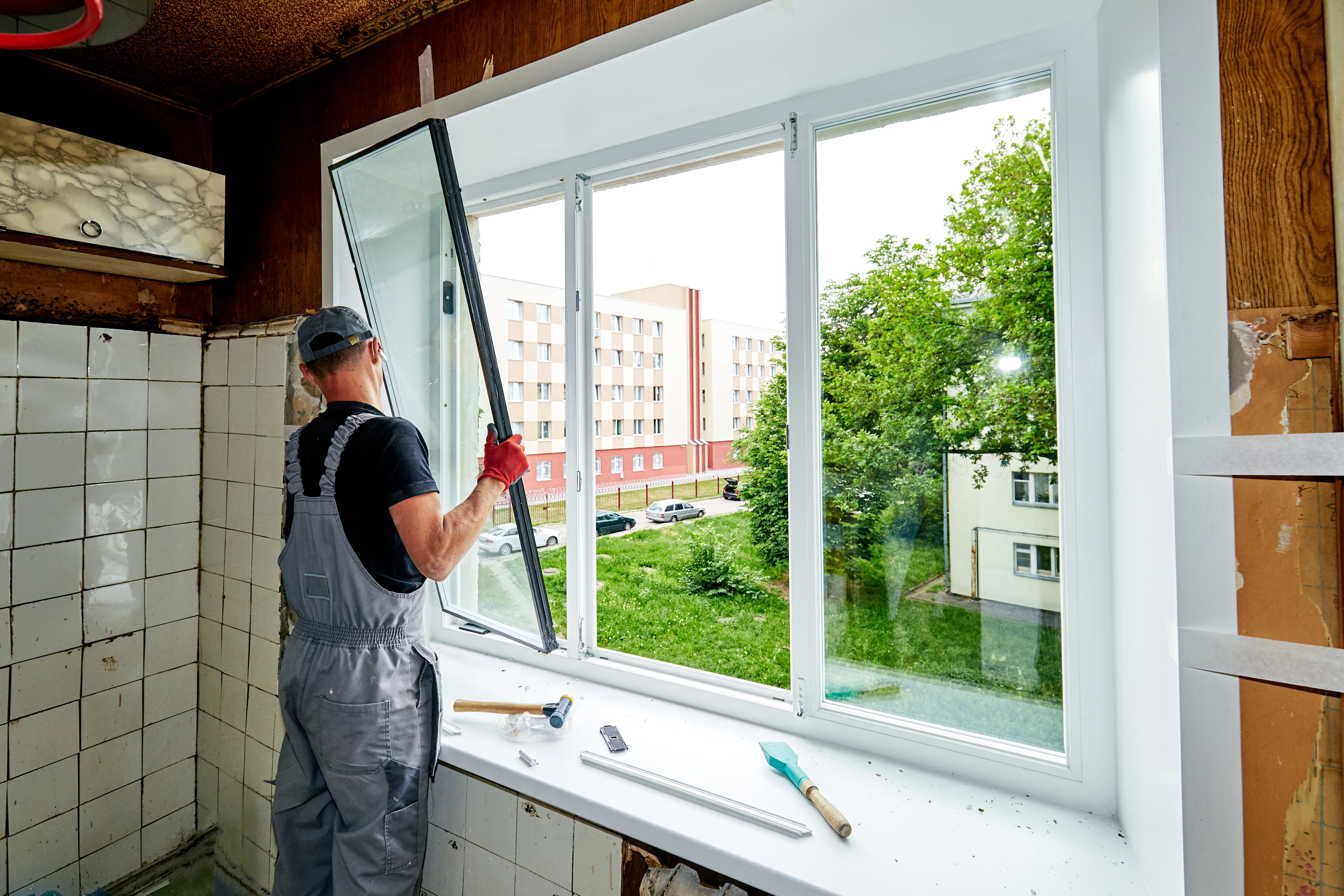 A worker installs a large window in a partially renovated room, with tools and a view of a green area outside.
