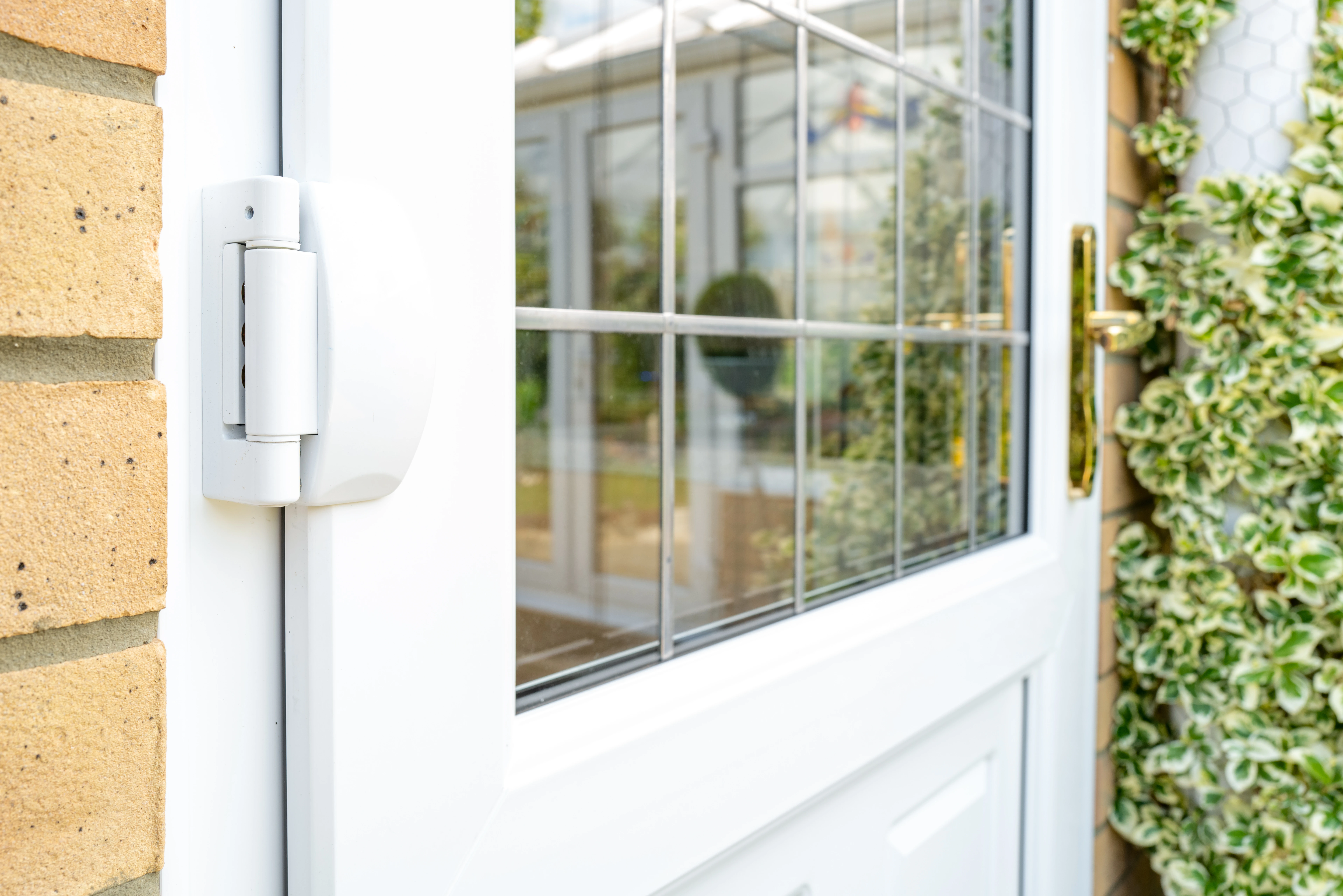 Close-up of a white door hinge and handle against a brick wall, with a glass panel showing a garden view and decorative greenery nearby.