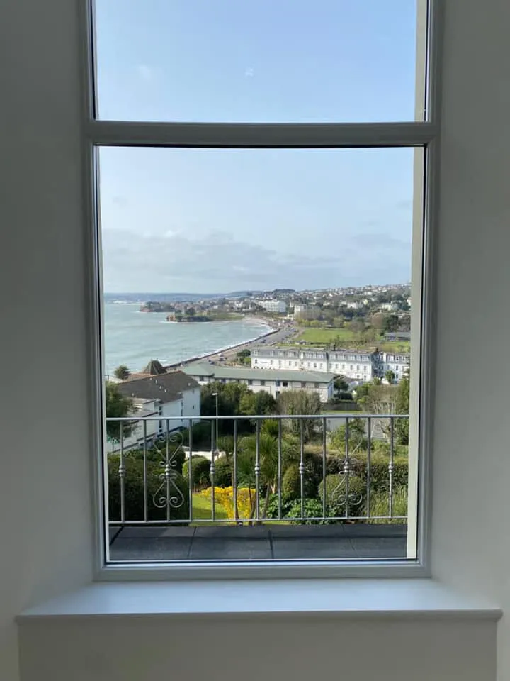 View through a large window showing a coastal landscape, with a beach, houses, and greenery in the foreground and background.