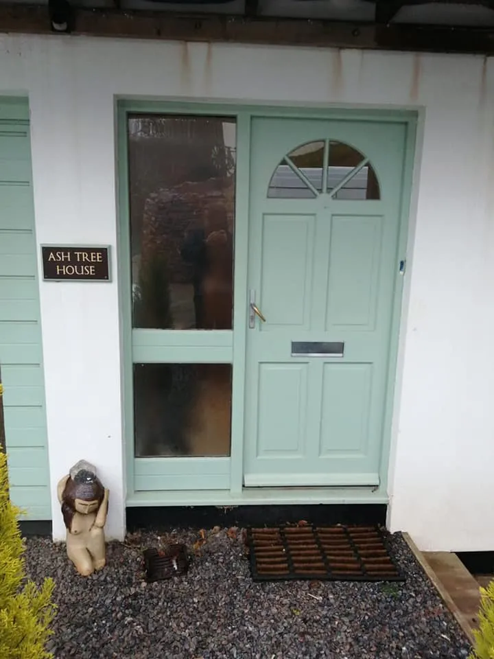 Mint green door with an arched window, a sign saying "Ash Tree House," and a decorative figure in front on gravel.