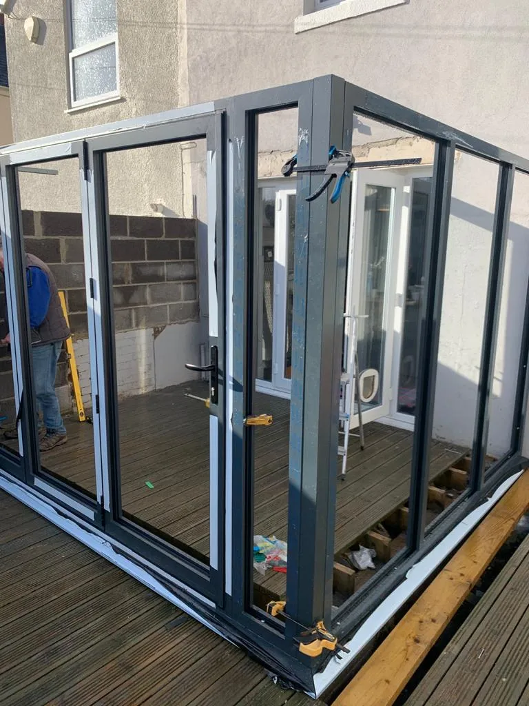 A partially constructed glass conservatory with metal frames, clamps, and wooden decking, showcasing tools and materials on the floor.