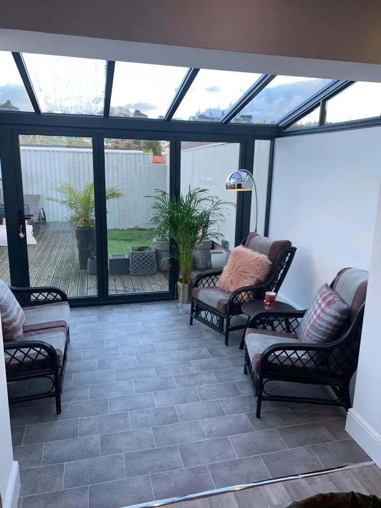 Bright sunroom with two chairs, a side table, and plants, featuring large glass doors opening to a decked garden area.