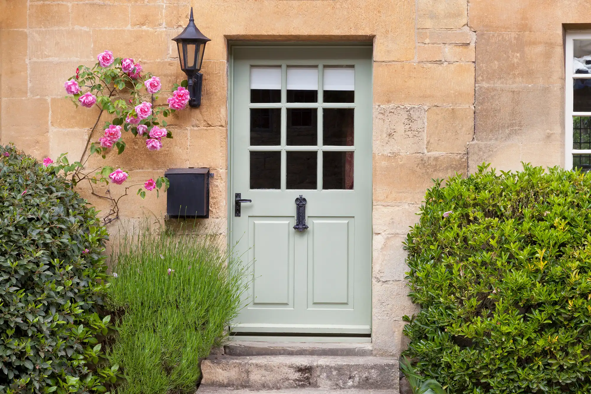 A charming green door with glass panes, surrounded by pink roses, a lamp, and lush greenery against a stone wall.